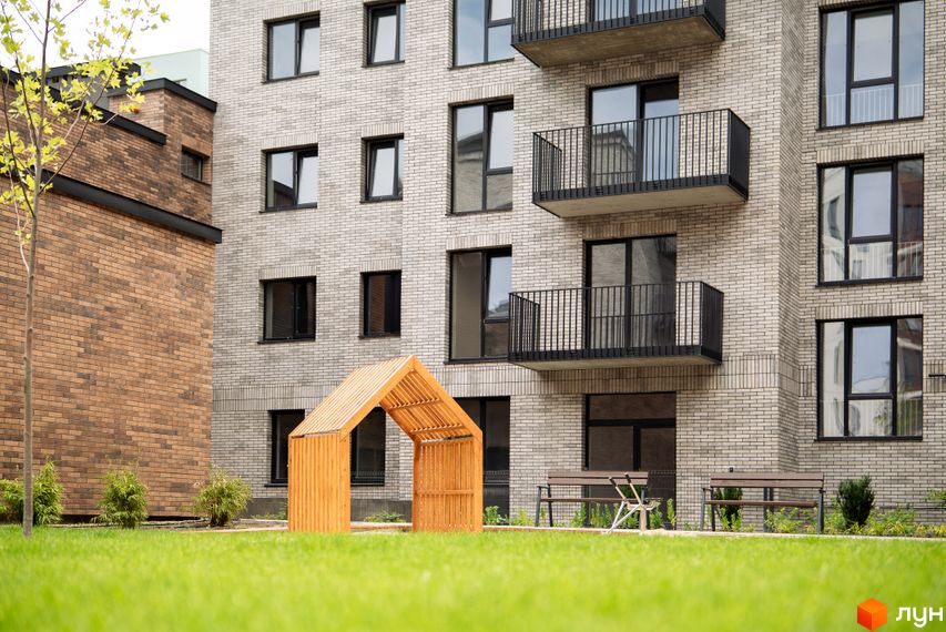 Будинок ЖК WAWEL з сірої цегли з балконами та прибудинкова територія з зеленим газоном та дерев'яною альтанкою.