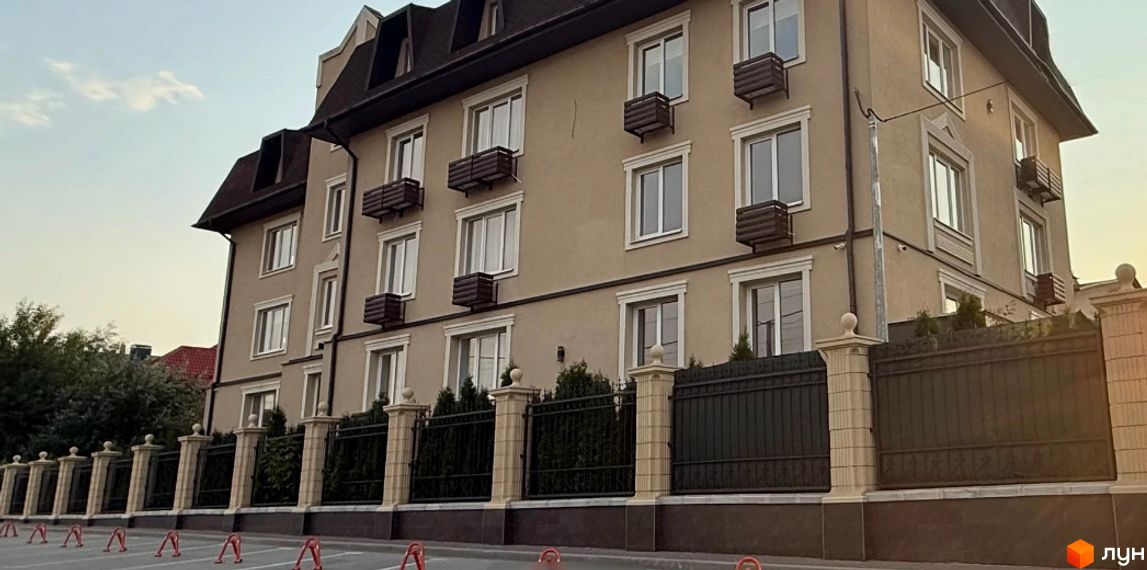 Зовнішній вигляд будинку ЖК Cappuccino House з вікнами та балкончиками, огородженого парканом з колонами. Навколо будинку зелені насадження.
