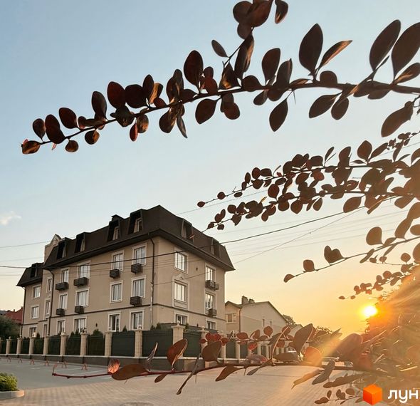 Новобудова ЖК Cappuccino House з п'ятьма поверхами та мансардою, вікнами з білими рамами та балконами. Навколо будинку розташована огорожа та зелені насадження.