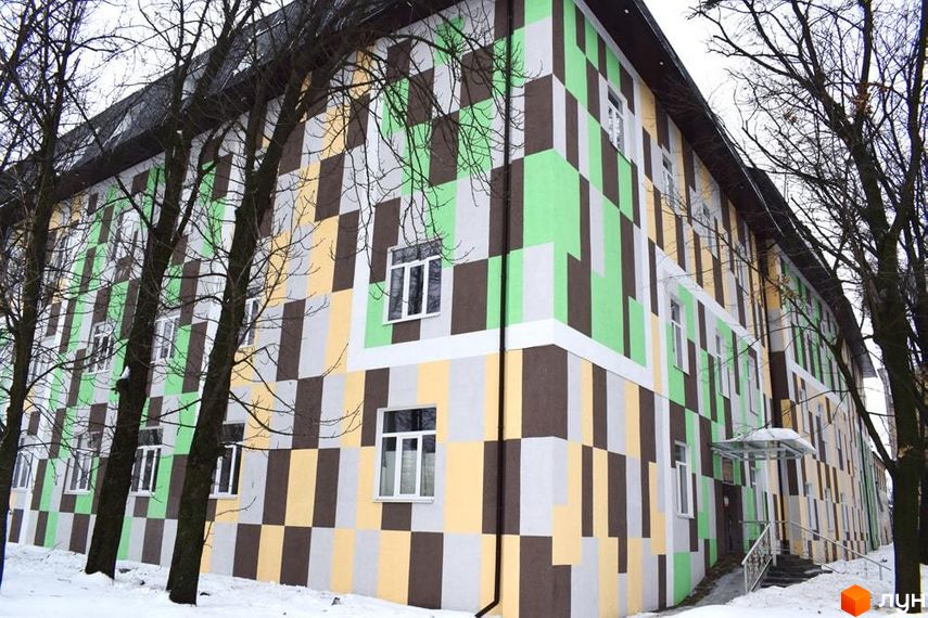 Житловий комплекс Нетеченський з фасадом, оздобленим прямокутними блоками зеленого, коричневого, жовтого та сірого кольорів. Навколо будинку сніг та дерева.