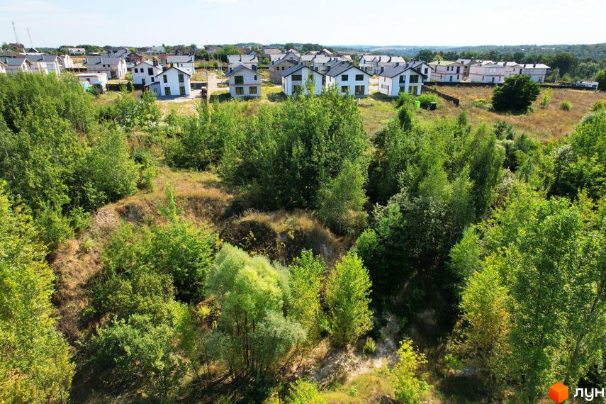 Котеджне містечко Панорама: білі двоповерхові будинки з чорними дахами на тлі зелених дерев та пагорбів. Навколо новобудов – природний ландшафт з лісом та травою.