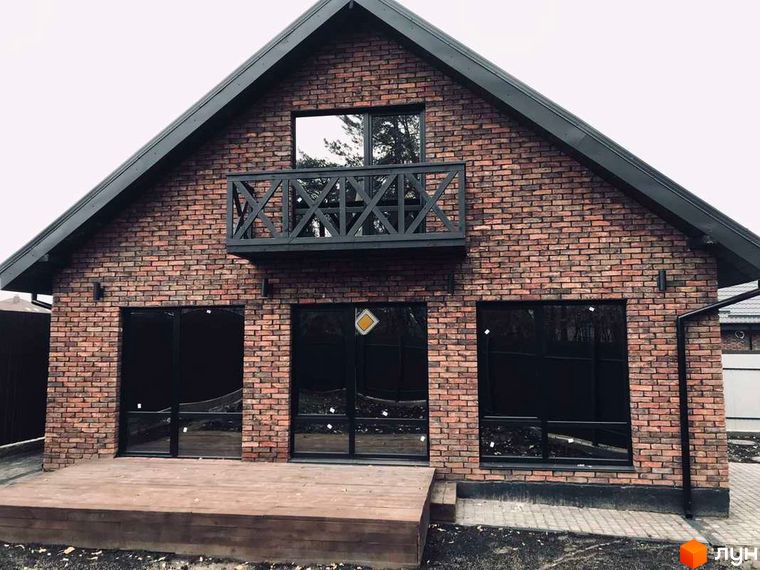 Котеджне містечко КМ Одноповерховий Київ - Дніпро. Фасад будинку з червоної цегли, великі чорні вікна, балкон з дерев'яною огорожею, дерев'яна тераса.