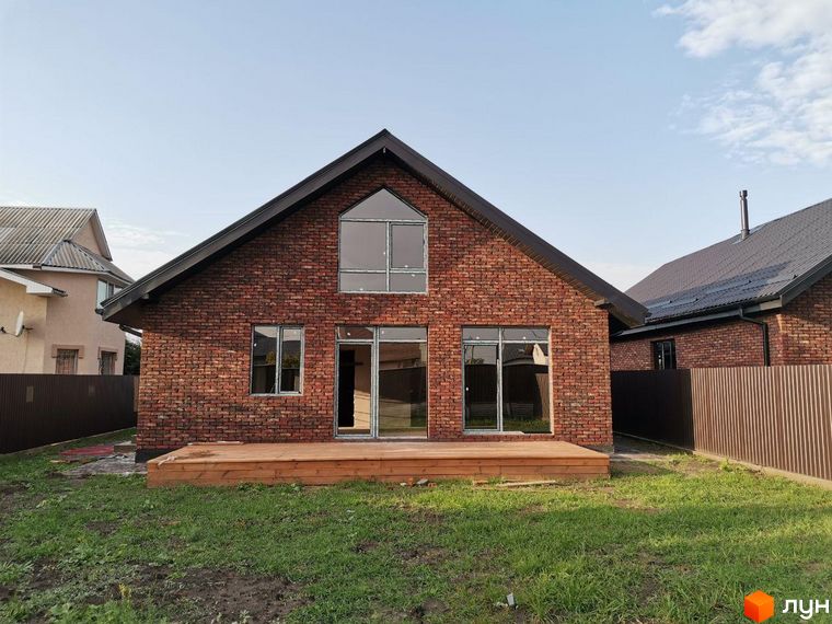 Котеджне містечко КМ Одноповерховий Київ - Дніпро. Фасад будинку з червоної цегли, великі вікна та дерев'яна тераса. Навколо будинку зелений газон.