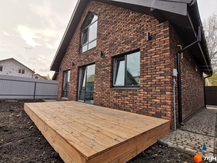 Котеджне містечко КМ Одноповерховий Київ - Дніпро. Зовнішній вигляд будинку з цегляним фасадом та дерев'яною терасою. Навколо будинку облаштована прибудинкова територія з бруківкою.