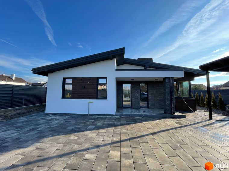 Новобудова: одноповерховий будинок з білими стінами, темним дахом та кам'яною обробкою, перед яким вимощена сіра бруківка.