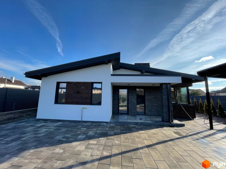 Сучасний одноповерховий будинок з білою штукатуркою, темним дахом та кам'яним оздобленням фасаду. Прибудинкова територія вимощена сірою плиткою.