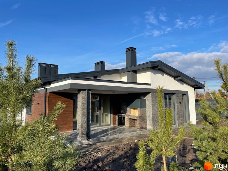 Одноповерховий будинок з темно-сірим дахом, білими та дерев'яними фасадами, критою терасою і зоною барбекю. Навколо молоді сосни та підготовлена ділянка на вул. Мазепи.