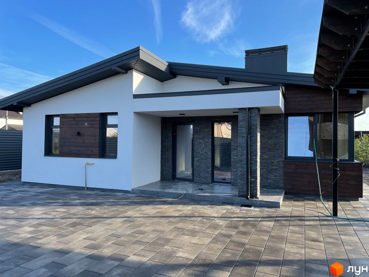 Одноповерховий будинок з комбінованим фасадом: білі стіни, дерев'яні панелі та кам'яне облицювання. Подвір'я вимощене бруківкою.