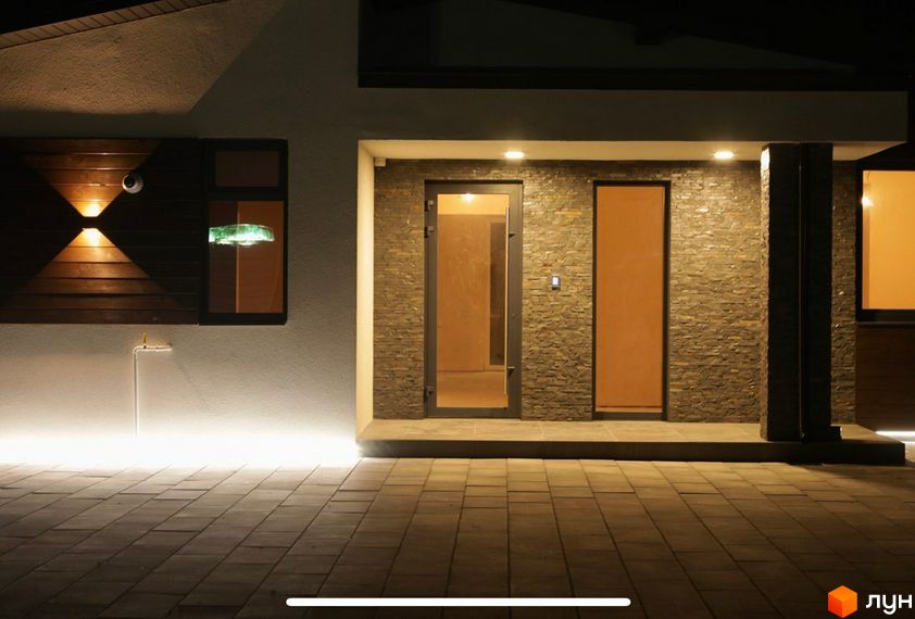 Житловий комплекс на вул. Мазепи: фасад будинку з кам'яним облицюванням, дерев'яною панеллю та освітленням. Прибудинкова територія вимощена плиткою з підсвічуванням.