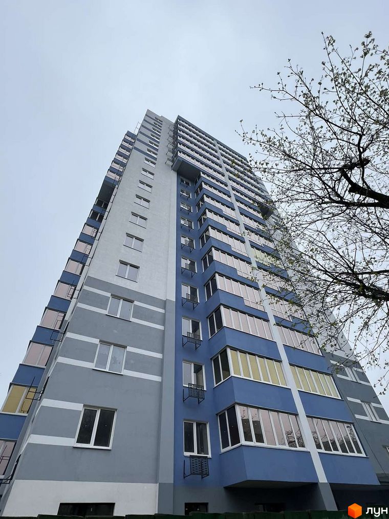 Зовнішній вигляд новобудови ЖК Diamond Hall з блакитно-сірим фасадом та скляними балконами. Будівельний проєкт розташований біля дерева з гілками.