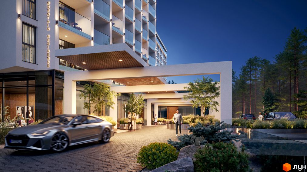 Апарт-комплекс Mountain Residence з підсвіченим фасадом та входом, оточений деревами та зеленими насадженнями. Перед комплексом їде сріблястий автомобіль.