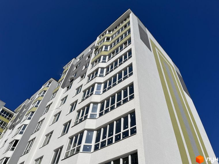 Висотна будівля житлового комплексу з великою кількістю вікон на фоні блакитного неба. Фасад будинку білого кольору з декоративними смугами.