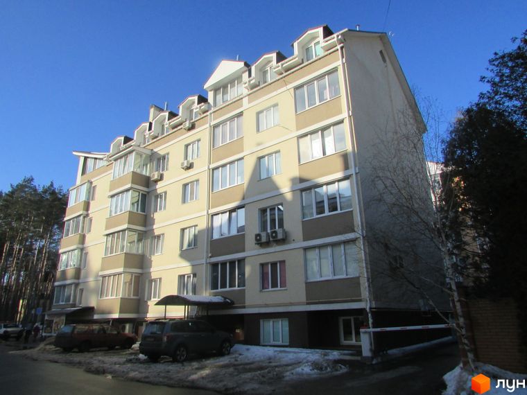Багатоповерховий житловий будинок світлого кольору з балконами та мансардними вікнами. На прибудинковій території припарковані автомобілі, видно дерева та сніг.