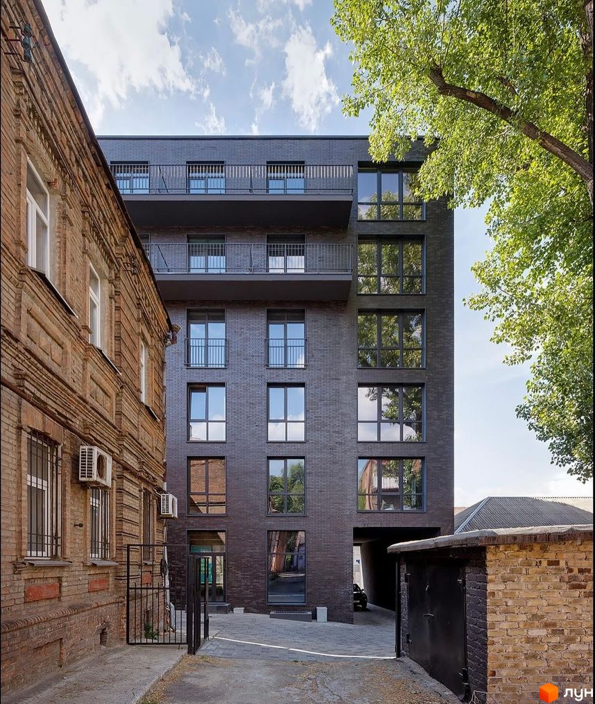 Темний цегляний багатоповерховий будинок Житлового комплексу ArtHof з великими вікнами та балконами. Поруч стара цегляна будівля та проїзд до паркінгу.