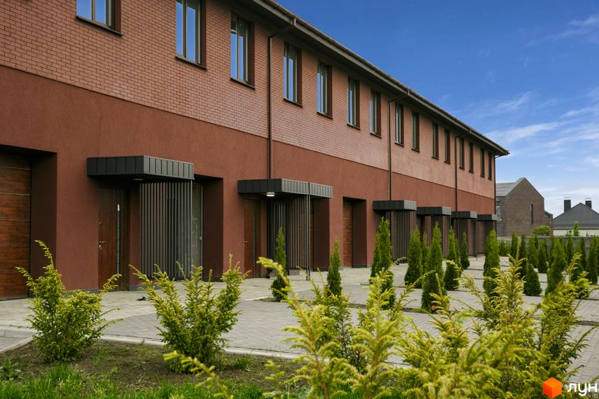 Зовнішній вигляд клубного будинку ЖК Обрій з червоної цегли та коричневої штукатурки, перед будинком висаджені туї та кущі.