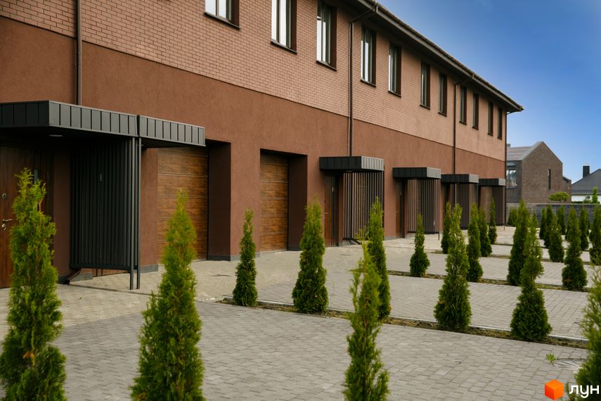 Ряд будинків Житлового комплексу клубного типу Обрій з коричневою цегляною кладкою та темно-коричневим фасадом. Перед будинками розташована вимощена територія з рядами зелених туй.