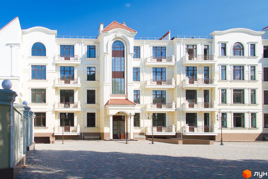 Житловий комплекс Клубний будинок || з фасадом у світлих тонах, балконами та великими вікнами. На передньому плані бруківка та ліхтарі.