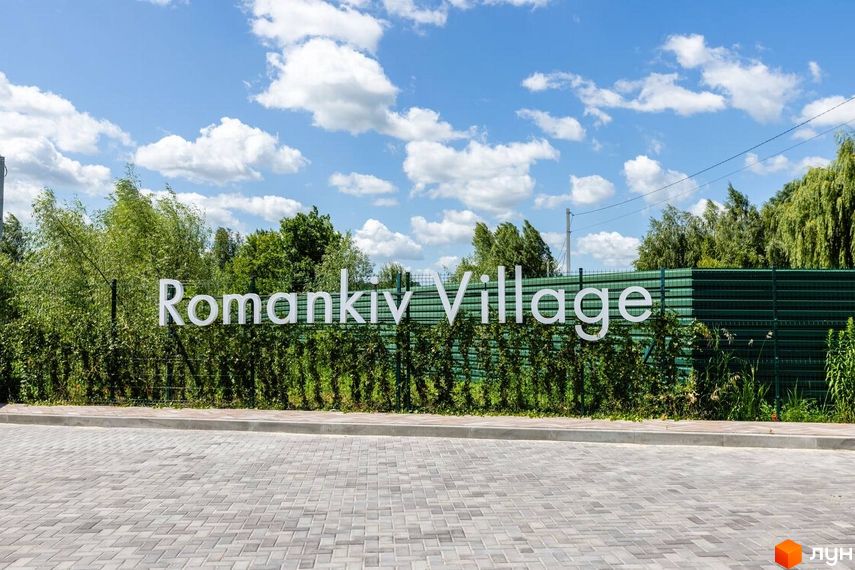 Котеджне містечко Romankiv Village. Білі об'ємні літери назви комплексу на фоні зеленого паркану та зелені. Перед комплексом вимощена сіра бруківка.