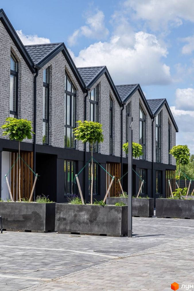 Котеджне містечко КМ Romankiv Village з сірої цегли під сірим дахом та облаштованою прибудинковою територією з деревами у великих бетонних вазонах.