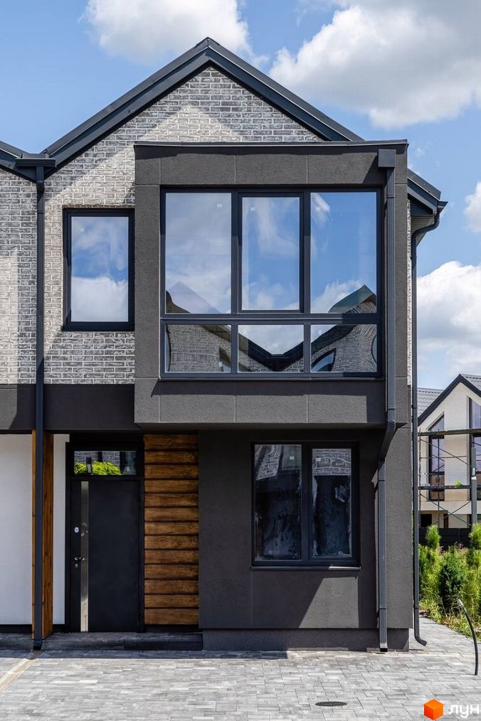 Зовнішній вигляд будинку в котеджному містечку КМ Romankiv Village з сірої цегли та темною штукатуркою, великими вікнами та дерев'яними елементами. На прибудинковій території видно бруківку та зелені насадження.