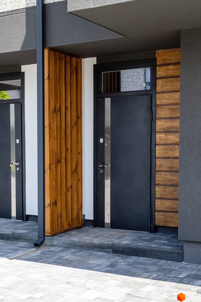 Зовнішній вигляд входу в котеджне містечко КМ Romankiv Village з темними дверима та дерев'яною обшивкою. Територія перед будинком вимощена плиткою.