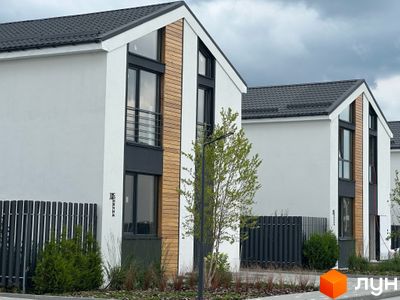 Котеджне містечко Gardd House Eco Village. Фасад будинку з білою штукатуркою, дерев'яними вставками та темними вікнами. Поруч паркан та зелені насадження.